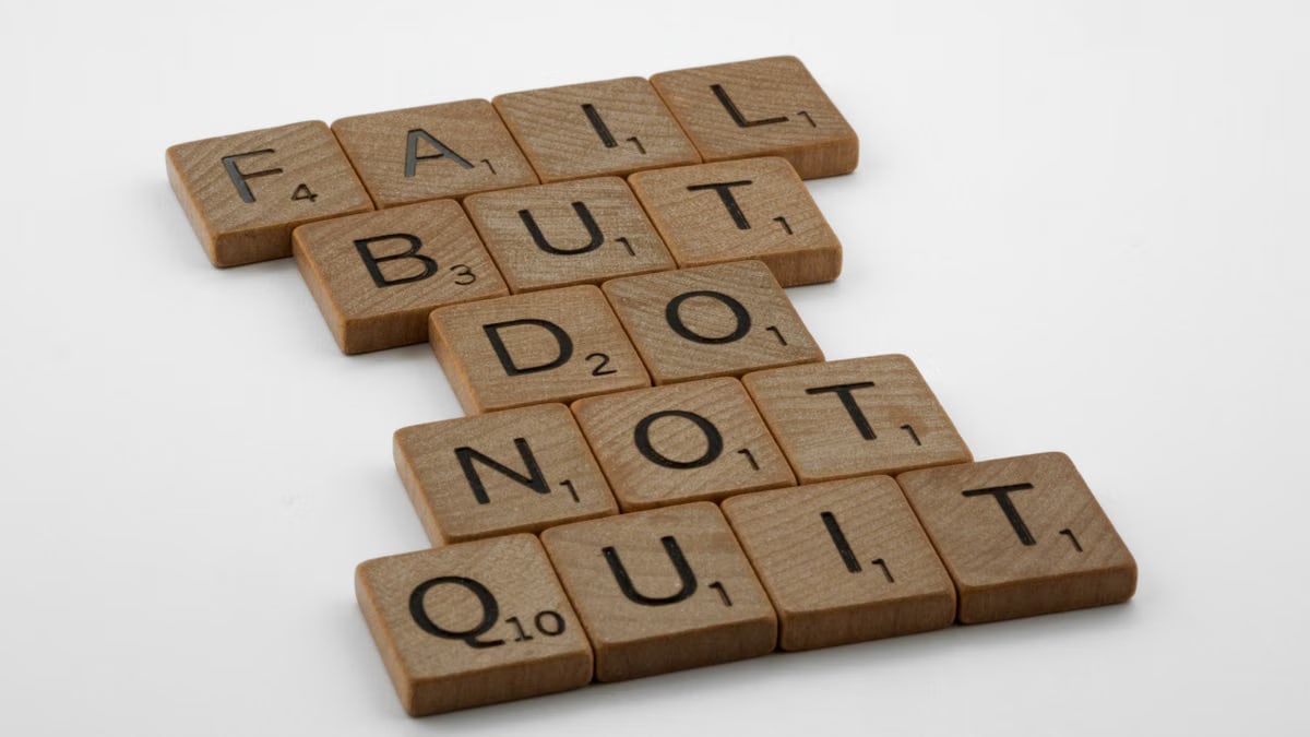 Close-up of Scrabble tiles forming the motivational phrase 'Fail but do not quit' on a white surface.